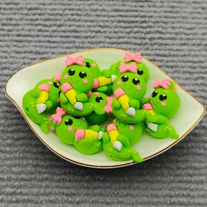 May include: A collection of green, cartoon-like figures with pink bows and pencils, arranged in a white and gold-rimmed dish. The figures have black eyes and are lying down. The dish is on a gray surface.