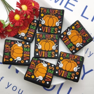 Op de afbeelding: Zes zwarte vierkante charmes met een pompoen en de tekst "Thankful Vibes" in kleurrijke letters. De charmes liggen op een wit oppervlak met een bloemenboeket.