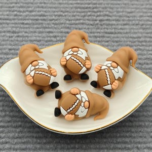 May include: Four brown gnome figurines with footballs, arranged on a white, leaf-shaped dish with a gold rim. The gnomes have tan hats, white beards, and black shoes. One gnome is lying on its back.