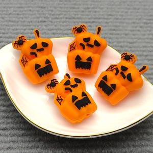 Op de afbeelding: Een verzameling oranje pompoenvormige decoraties met zwarte gezichten en spinnenaccenten, gerangschikt op een witte plaat met een gouden rand. De pompoenen zijn gestapeld en hebben een cartoonachtig ontwerp, perfect voor Halloween.