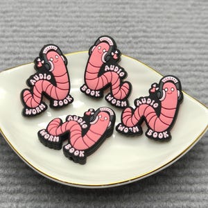 May include: A collection of pink worm-shaped charms with the words "Audio Book Worm" printed on them. Each charm has a black outline and is wearing headphones. The charms are arranged on a white, gold-rimmed dish.
