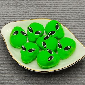 Puede incluir: Un plato blanco en forma de hoja con borde dorado contiene siete objetos verdes con forma de cabeza de alienígena. Cada alienígena tiene grandes ojos negros. El plato está sobre una superficie gris texturizada.