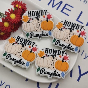 Op de afbeelding: Vier witte en oranje pompoen-vormige ornamenten met de tekst "Howdy Pumpkin" op elk. De ornamenten zijn versierd met herfstbladeren en bloemen.