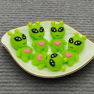 Puede incluir: Un conjunto de figuras de alienígenas verdes con ojos negros, pestañas y corazones rosas en el pecho. Los alienígenas tienen antenas amarillas en forma de estrella, dispuestas en un plato blanco con borde dorado.