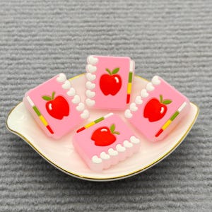 May include: Four pink, rectangular charms shaped like notebooks, each featuring a red apple illustration. The charms are arranged on a white and gold-rimmed dish. The background is a gray, textured surface.