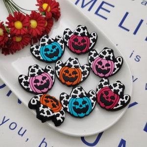 Op de afbeelding: Acht kleurrijke Halloween-pompoencharms met een zwart-witte koe-print ontwerp. Elke charme heeft een pompoen met een jack-o'-lantern gezicht dat een cowboyhoed draagt.