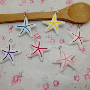 May include: Six white starfish charms with different colored lines. The starfish are hanging from silver jump rings. The charms are on a white background with pink flowers.