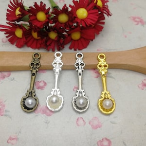 May include: Four antique silver spoon charms with a pearl on the end. The charms are arranged on a wooden surface.
