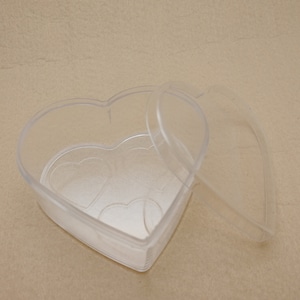 Op de afbeelding: Een doorzichtige, hartvormige plastic doos met een bijpassende deksel. De doos is open, waardoor de binnenkant zichtbaar is, en staat op een neutrale ondergrond. Het ontwerp bevat hartvormige details op de basis, wat wijst op een thema van liefde of romantiek.