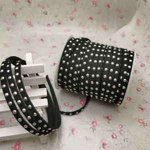 May include: A spool of black leather cord with silver studs. The cord is wrapped around a white spool and there is a length of cord lying on a white surface.