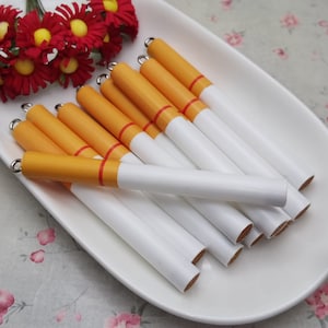 May include: A collection of white and orange cigarette-shaped pens arranged on a white oval plate. The pens have a yellow-orange tip and a red band. A bouquet of red and yellow flowers sits above the pens.