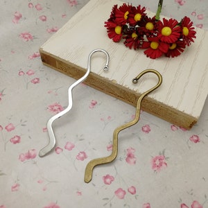 May include: Two silver and gold bookmark pendants with a wavy design. The bookmarks are on a white surface with a floral pattern.