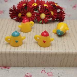 Puede incluir: Cuatro teteras amarillas en miniatura con detalles azules y rojos. Las teteras tienen un diseño de corazón en la parte delantera y una tapa con una parte superior azul o roja.