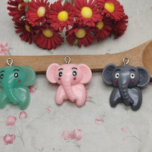 Könnte beinhalten: Drei kleine Elefanten-Anhänger in verschiedenen Farben. Die Elefanten sind grün, rosa und grau. Jeder Elefant hat eine silberne Schlaufe zum Aufhängen.