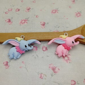May include: Two small, colourful elephant charms. One is light blue with pink and yellow accents, the other is pink with blue and yellow accents. Both have a silver-coloured loop at the top for hanging.