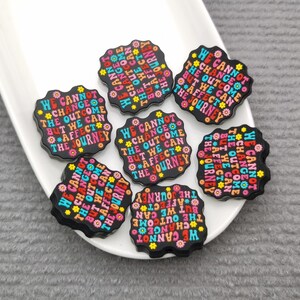 May include: Seven black round resin flatback cabochons with a colorful floral design and the text "We cannot change the outcome but we can affect the journey".