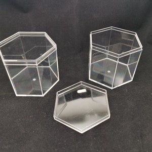 May include: Three clear, hexagonal acrylic boxes with lids. Two boxes are upright, and one lid is separate. The boxes are transparent, allowing visibility of the contents. The design is geometric and modern, suitable for storage or display.