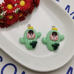 以下が含まれることがあります： 笑顔の顔、茶色の髪、黄色の花のアクセントが付いた緑色のサボテンチャーム2つ。チャームには、吊り下げ用の銀色のループが付いています。