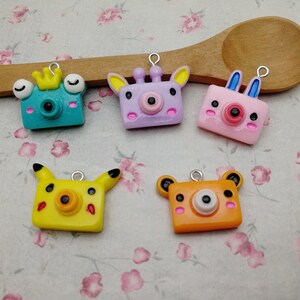 May include: Six colorful camera charms with animal faces. The charms are made of polymer clay and have a loop for attaching to a keychain or bag. The animals include a frog, a giraffe, a bunny, a Pikachu, and a bear.