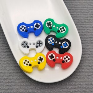 May include: Six colorful plastic game controller charms in blue, green, white, black, yellow, and red. The charms are arranged in a circle on a white plate.