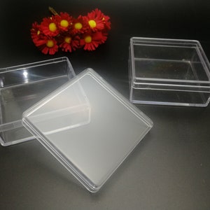 Könnte beinhalten: Transparente, quadratische Kunststoffboxen mit Deckeln, ideal für kleine Gegenstände. Eine Box ist geöffnet, der Deckel daneben. Eine andere Box ist geschlossen. Rote und gelbe Blumen sind im Hintergrund. Diese Boxen eignen sich für Bastelarbeiten, Aufbewahrung oder Geschenke.