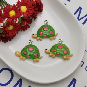 Puede incluir: Tres encantos de tortuga verde con lunares rosas y lazos plateados. Las tortugas están sobre una superficie blanca.