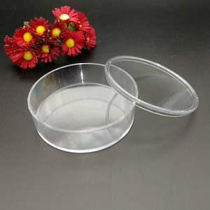 Op de afbeelding: Een ronde, transparante plastic container met een deksel. De container is leeg en het deksel is eraf. De container staat op een zwart oppervlak.