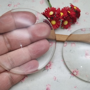 May include: Two clear glass magnifying lenses, one held in a hand and the other on a wooden spoon. The lenses are round and have a slight convex shape.