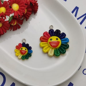 Puede incluir: Dos encantos de flores coloridos con una cara sonriente. El encanto más grande es de color arcoíris con un centro amarillo y una sonrisa roja. El encanto más pequeño es del mismo diseño pero de tamaño más pequeño.