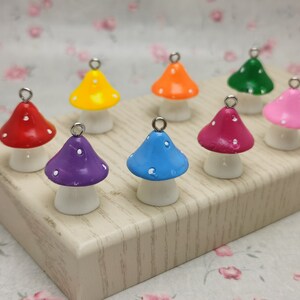 May include: Assortment of eight colourful mushroom charms with white polka dots. Each charm has a silver loop for hanging. The charms are arranged on a light wood surface.