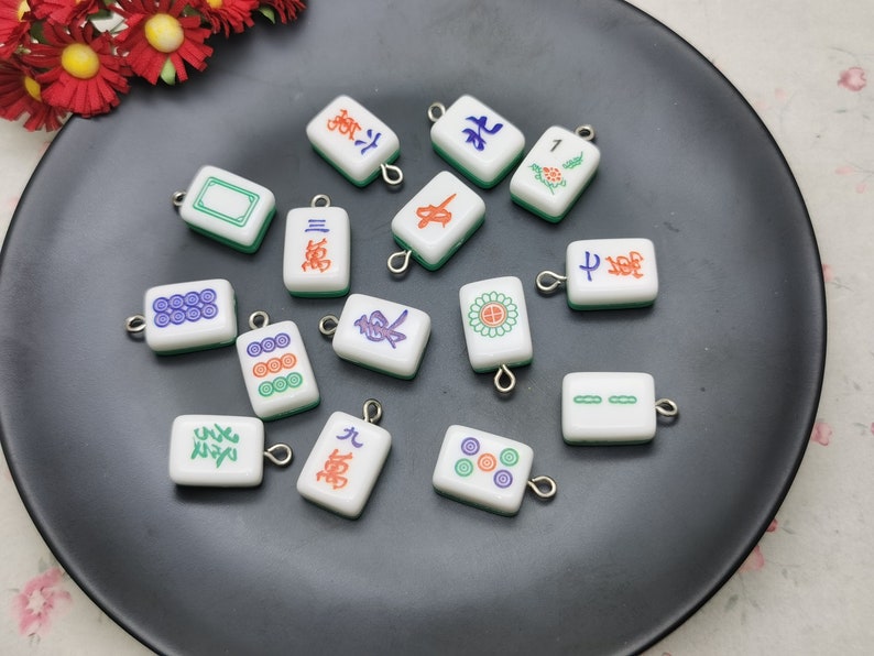 May include: A collection of small, white Mahjong tile charms with various symbols and characters in red, green, and blue. Each tile has a green border and a silver-colored loop for attaching to jewelry or other crafts. The charms are displayed on a black plate.