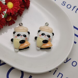 May include: Two white panda bear charms with black eyes and a black nose. Each panda is wearing a cream colored hat and holding a yellow and orange slice of fruit. The charms are on a white plate.