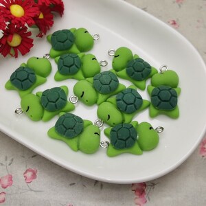 Puede incluir: Una colección de pequeños dijes de tortuga verde con detalles de caparazón verde oscuro, dispuestos sobre un plato ovalado blanco. Cada dije tiene un lazo plateado para sujetarlo a joyas u otras manualidades. Flores de papel rojas con centros amarillos en el fondo.