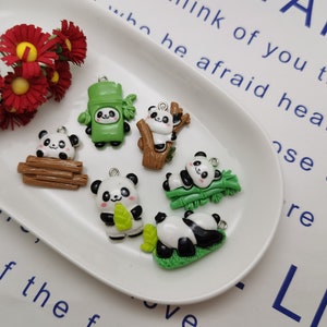May include: Six panda charm pendants on a white plate. The charms are made of polymer clay and feature different panda designs, including a panda holding bamboo, a panda sitting on a log, and a panda in a bamboo forest.
