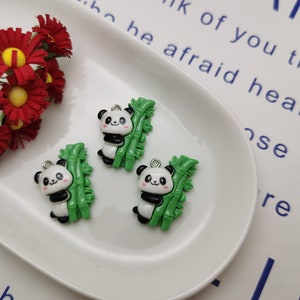 May include: Three panda charms with green bamboo stalks. The pandas are black and white with small pink noses and eyes. The charms are made of plastic and have a silver loop for hanging.