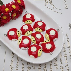May include: A selection of red mushroom charms with white spots and yellow faces, presented on a white oval dish. Each charm has a silver loop for attaching. Red flowers with yellow centres are also visible in the image.