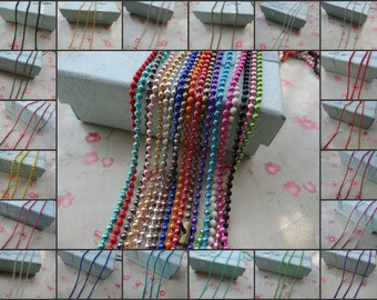 May include: A collection of colorful ball chain necklaces in various colors, including black, gold, red, and blue. The necklaces are displayed in front of small, light blue gift boxes. The background has a floral pattern.