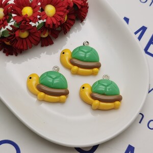 Puede incluir: Tres dijes de tortuga amarillos con caparazones verdes y detalles marrones. Las tortugas tienen anillos de salto plateados para sujetar a joyas u otras manualidades.