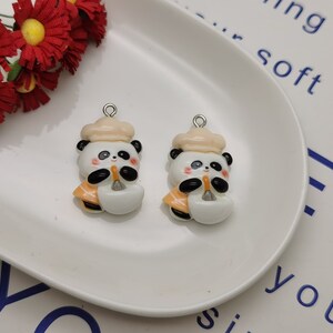 May include: Two white ceramic panda bear charms with orange accents. Each panda is wearing a cream-colored chef's hat and holding a small orange and white object. The charms have silver jump rings for attaching to jewelry or other crafts.