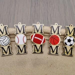 Könnte beinhalten: Fünf Holzornamente mit dem Wort "MOM" in schwarzen Buchstaben. Jedes Ornament zeigt einen anderen Sportball: Volleyball, Baseball, Fußball, Basketball und Fußball.