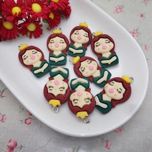 May include: Eight green and red resin charms of a girl with a crown, closed eyes, and a pink blush. Each charm has a gold loop for hanging.