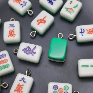 May include: A collection of small, rectangular Mahjong tile charms. The charms are white with green edges and feature various symbols and characters in red, blue, and purple. Each charm has a silver loop for attaching to jewelry or other crafts.