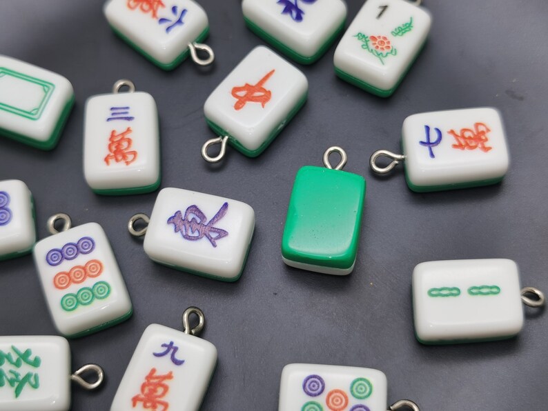 May include: A collection of small, rectangular Mahjong tile charms. The charms are white with green edges and feature various symbols and characters in red, blue, and purple. Each charm has a silver loop for attaching to jewelry or other crafts.