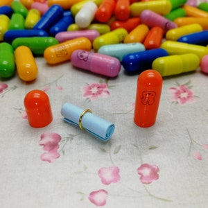 May include: A close-up of two orange plastic capsules with a rolled up blue paper note and a gold ring. The capsules have a white design on them. The capsules are sitting on a white surface with a floral pattern.