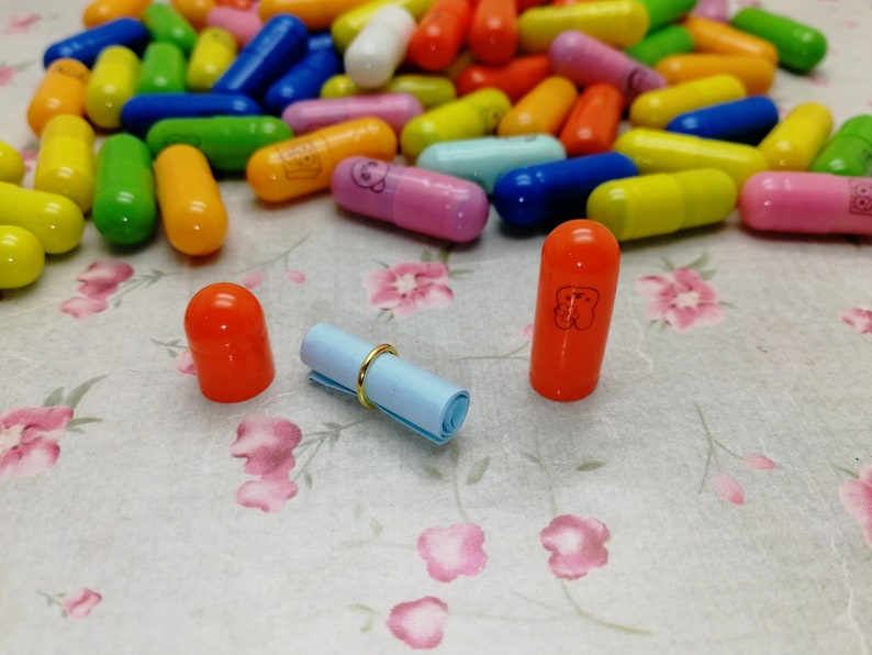 May include: A close-up of two orange plastic capsules with a rolled up blue paper note and a gold ring. The capsules have a white design on them. The capsules are sitting on a white surface with a floral pattern.
