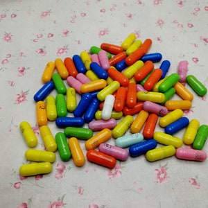 May include: A large pile of colorful capsules, mostly in shades of yellow, orange, green, blue, and pink. There is one white capsule in the center of the pile.