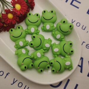 May include: A collection of nine green frog-shaped charms with white eyes and smiling faces. The charms are made of a soft, pliable material and are perfect for adding a whimsical touch to crafts and jewelry.