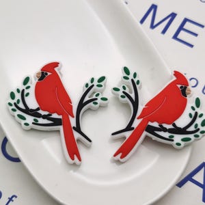 Può includere: Due ciondoli a forma di cardinale rosso con foglie verdi su sfondo bianco. Gli uccelli sono appollaiati su rami e hanno occhi e becchi neri.