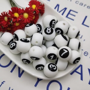 Puede incluir: Un bol blanco lleno de cuentas de plástico blanco con números negros en ellas. Los números van del 1 al 8. El bol está sobre una superficie blanca con texto azul.