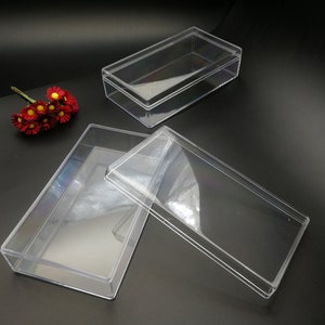 May include: Transparent rectangular plastic boxes with lids, suitable for storage or display. One box is open, lid beside it. Another box is closed. A small bouquet of red and yellow flowers is in the background.